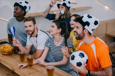 multicultural friends in soccer ball hats celebrating, gesturing by hands and watching football match at bar  clipart