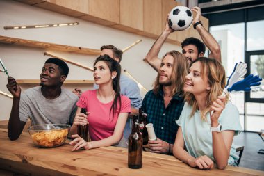multicultural friends with football ball and hand clappers during watch of soccer match at bar  clipart