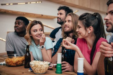 multiethnic group of friends watching football match at bar with fan horns, chips, popcorn, beer and soccer ball  clipart