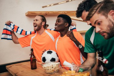 çok kültürlü futbol hayranları t-shirt ve farklı t-shirt yakınındaki futbol seyretmek sırasında oturan üzgün onların arkadaşları ise topu ile zaferini kutluyor eşarp maç, turuncu gülümseyen bar