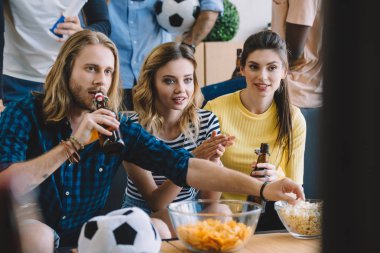 cropped shot of friends with soccer ball, beer bottles, bowls with popcorn and chips watching football match at home clipart