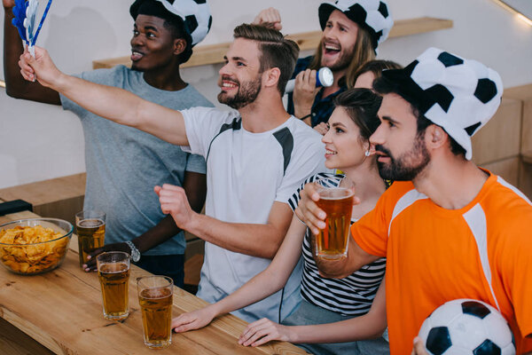 улыбающиеся мультикультурные друзья в футбольных бейсболках празднующие, пьющие пиво и смотрящие футбольный матч в баре
 