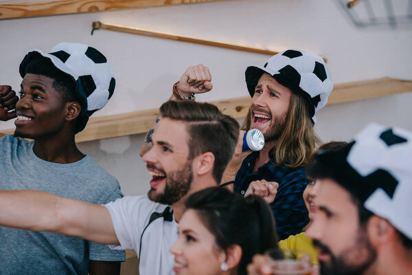 happy multicultural friends in soccer ball hats celebrating, gesturing by hands and watching football match at bar 
