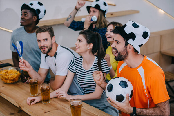 мультикультурные друзья в футбольных бейсболках празднуют, жестикулируют руками и смотрят футбольный матч в баре
 