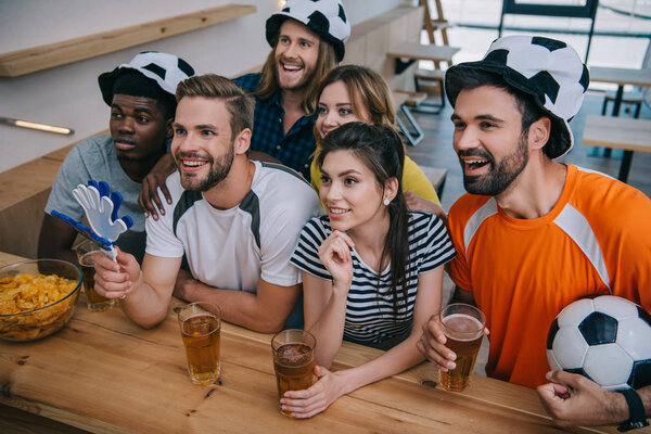 счастливая мультикультурная группа друзей в футбольных кепках, пьющих пиво и смотрящих футбольный матч в баре
 