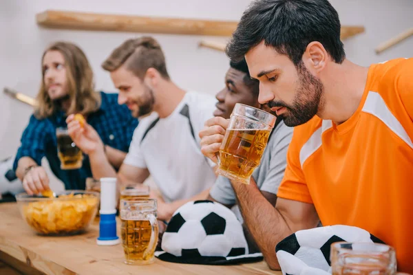 side view of frustrated young man in orange t-shirt drinking beer while his friends sitting behind at bar counter with ball hats, chips, fan horn and beer during watch of soccer match