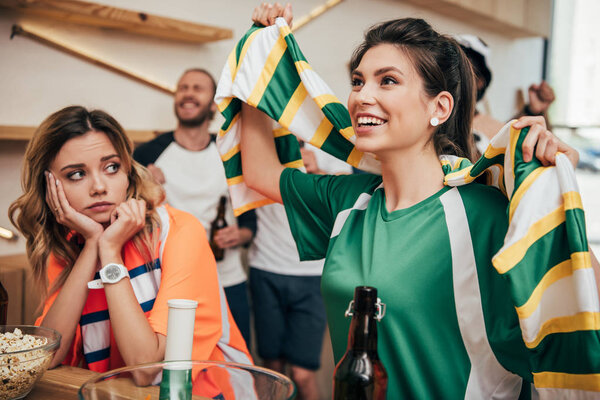 счастливая молодая женщина в зеленой футболке с веером и шарфе, празднующая в то время как ее расстроенная подруга в оранжевой футболке сидит во время просмотра футбольного матча в баре
 