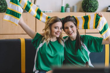 smiling female football fans in green t-shirts and scarf celebrating during watch of soccer match at home clipart