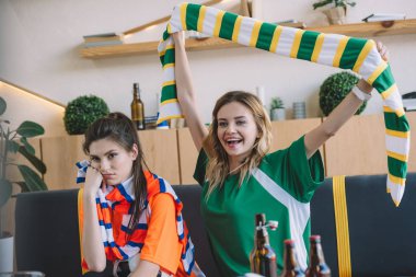 gülümseyen kadın yeşil fan t-shirt kutluyor zafer ve üzerinde kafa Turuncu tişört yakınındaki sırasında futbol izle kanepede oturan üzgün onun kız arkadaşı ise maç eşarp evde tutan