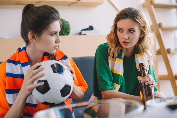 two female football fans in different fan t-shirts with ball and beer bottle looking at each other at home