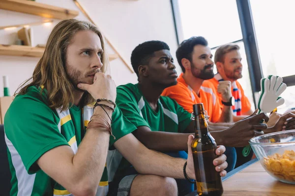 focused group of multicultural male friends in different fan t-shirts watching soccer match at home