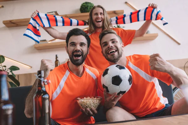兴奋的年轻男性足球爱好者在橙色 T恤与围巾 球和爆米花庆祝和观看足球比赛在家里 — 图库照片