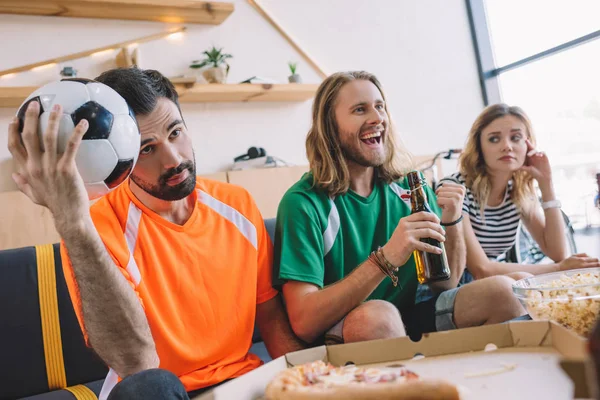 緑ファン シャツはサッカーの観戦中にソファの近くに座って彼の怒っている友人が自宅と一致を祝う幸せな若い男  — 無料ストックフォト