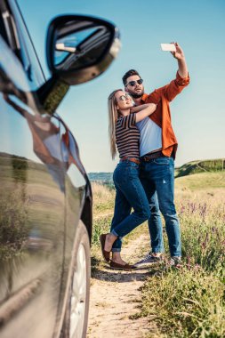 stylish man in sunglasses taking selfie on smartphone with smiling girlfriend near car on rural meadow  clipart