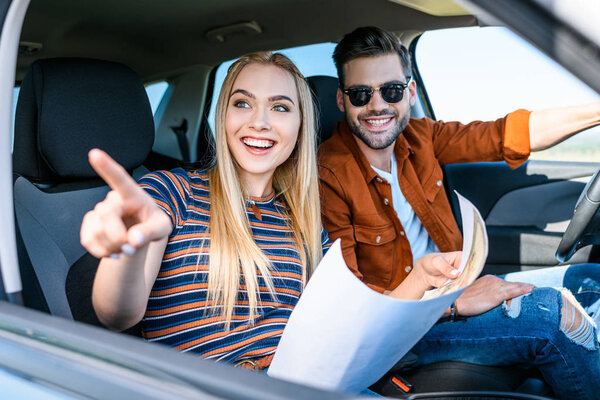 young woman with map in hand pointing by finger to smiling boyfriend sitting behind car wheel