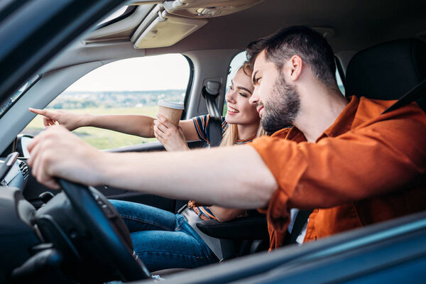 smiling woman with cup of coffee pointing by finger to boyfriend sitting behind car wheel