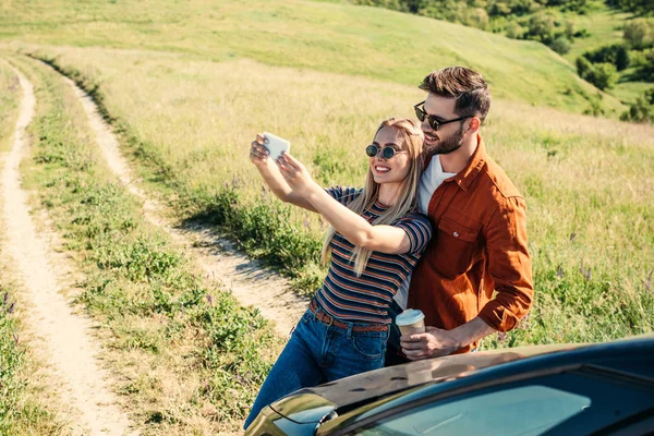 Stylowy Para Okulary Filiżanki Kawy Biorąc Selfie Smartfon Pobliżu Samochodu — Zdjęcie stockowe