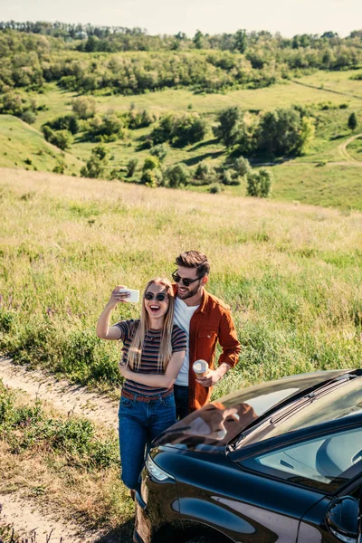高角度的微笑时尚情侣在太阳镜与咖啡杯自拍在农村草地上的智能手机附近汽车 — 免费的图库照片