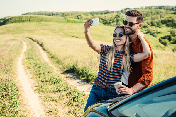 smiling stylish couple in sunglasses with coffee cup taking selfie on smartphone near car on rural meadow 