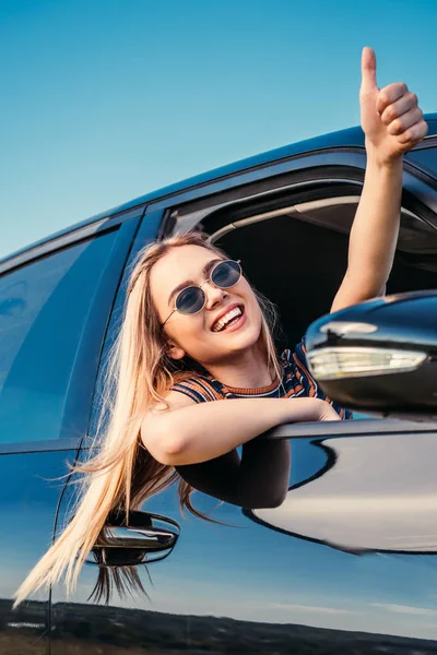 微笑时尚的妇女在太阳镜倾斜从汽车窗口和做拇指向上手势 — 图库照片