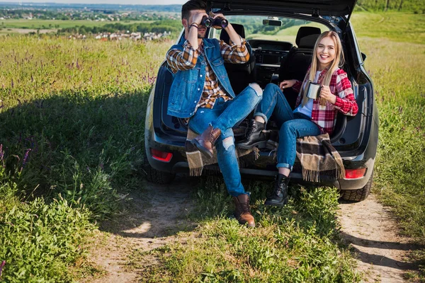 男性游客看着望远镜 而他的微笑的女朋友坐在附近的汽车树干在农村领域的咖啡杯 — 图库照片