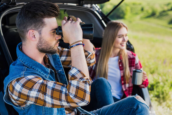 вид сбоку турист смотрит через бинокль в то время как его улыбающаяся подруга сидит рядом с чашкой кофе на багажнике автомобиля
 