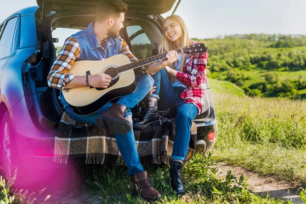 Young Male Traveler Playing Acoustic Guitar Smiling Girlfriend Car Trunk — Stock Photo, Image