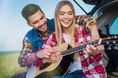 young man teaching smiling girlfriend to play on acoustic guitar while they sitting on car trunk  clipart