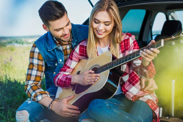 male tourist teaching smiling girlfriend to play on acoustic guitar on car trunk in rural field
