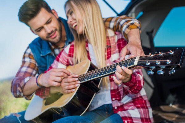 smiling man teaching smiling girlfriend to play on acoustic guitar on car trunk 