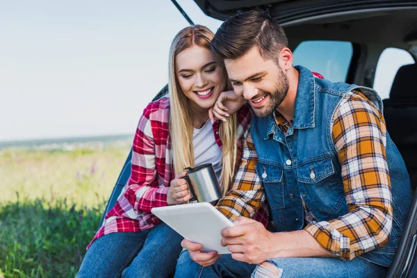 微笑的人使用数码片 而他的女朋友坐在附近的汽车树干在农村领域的咖啡杯 — 图库照片