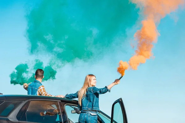 young couple standing on car and holding green and orange smoke bombs