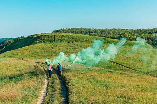 田舎の牧草地の緑の煙爆弾を抱いた若い夫婦の遠景  — 無料ストックフォト