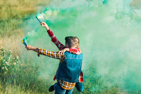 rear view of man holding girlfriend and green smoke bombs in field 