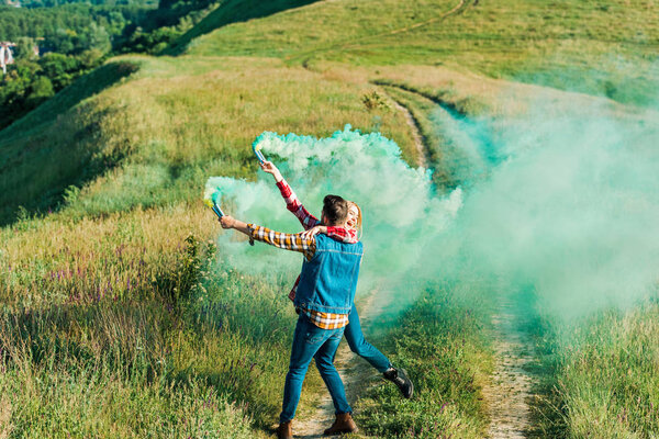 back view of man holding girlfriend and green smoke bombs on rural meadow