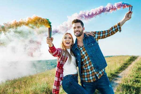 田舎の牧草地にカラフルな煙爆弾を保持している笑顔のカップル — ストック写真