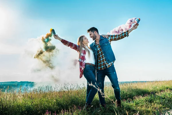 年轻夫妇看着对方 并持有彩色烟雾炸弹在农村草地上 — 图库照片