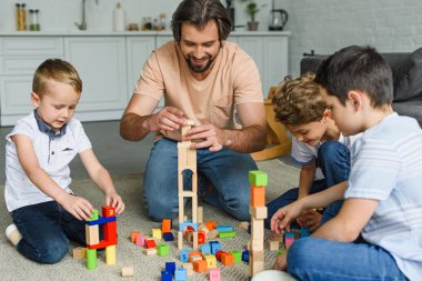 cheerful father and kids playing with wooden blocks together on floor at home clipart