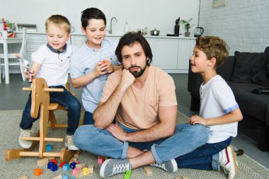 stressed father sitting on floor and sons playing around in room clipart