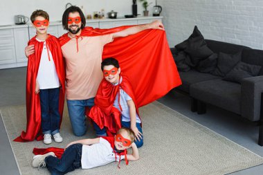 smiling father and sons in red superhero costumes at home clipart