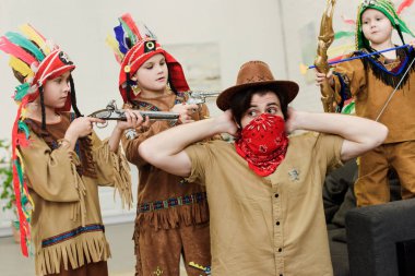 father in hat and bandana and little sons in indigenous costumes with toys playing together at home clipart