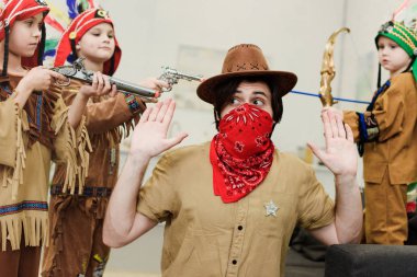 father in hat and bandana and little sons in indigenous costumes with toys playing together at home clipart