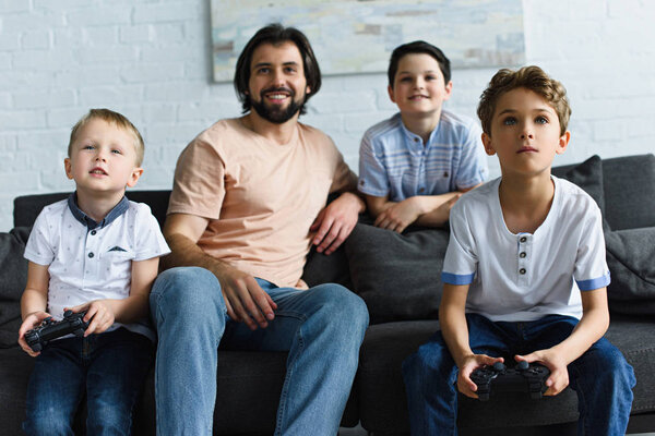 smiling father looking at little sons sitting on sofa and playing video games together at home