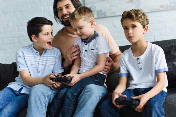 smiling father looking at little sons sitting on sofa and playing video games together at home