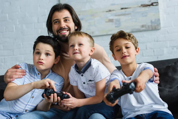 Sonriente Padre Mirando Los Pequeños Hijos Sentados Sofá Jugando Videojuegos —  Fotos de Stock