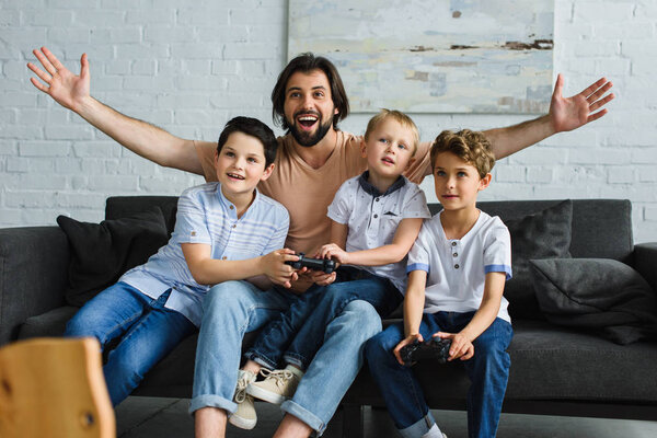 excited father and little sons sitting on sofa and playing video games together at home