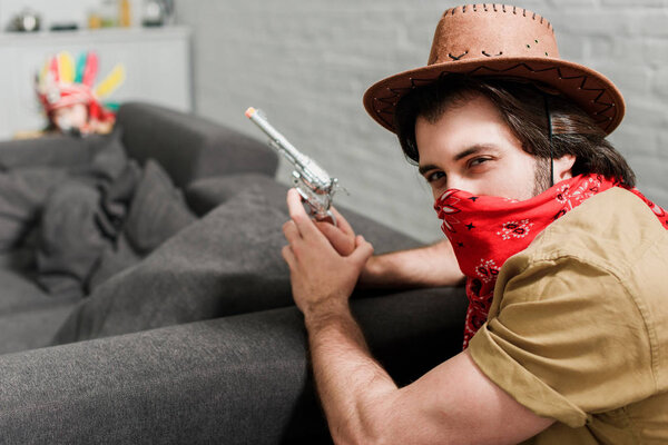 side view of man in red bandana and cowboy hat with toy gun at sofa at home