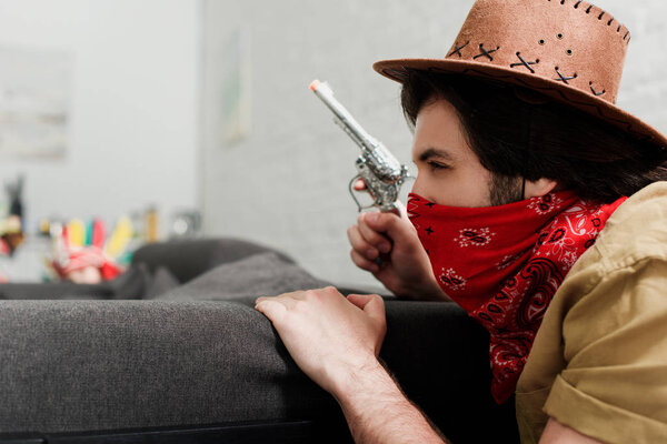 side view of man in red bandana and cowboy hat with toy gun at sofa at home