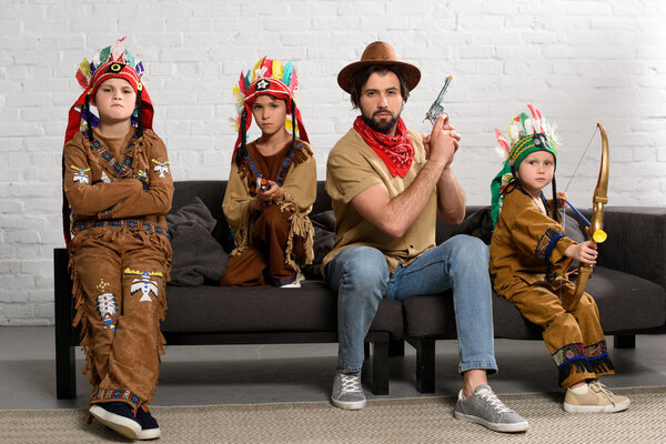 father in hat and red bandana sitting on sofa with little sons in indigenous costumes at home