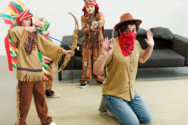 father in hat and bandana and little sons in indigenous costumes with toys playing together at home